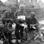 Pfc Edmund Dill (Cumberland Md) opens the Christmas package received from his wife. His buddies share the treat. 18 November, 1944 left Pfc Carl Anker (Brooklyn NY) right, Sgt Ted Bailey (Isaban W.Va) 30-ID(scaled)