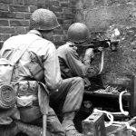 Pfc Victor Henry (Pontotoc, Miss), fires his machine gun through a hole in a wall, at Germans in a barn 300 yards away, beyond Kohlscheid, Germany 16 October, 1944 (scaled).