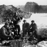 The crew of a Self-Propelled 105-MM Howitzer keep warm around a fire, between the fire missions near Les Tailles, Belgium