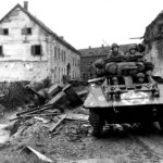 An M8 reconnaissance armored car of the 30th Infantry Division moves through the streets of Kinzweiler, Germany, past an abandoned German StuG, November 21, 1944-(scaled)